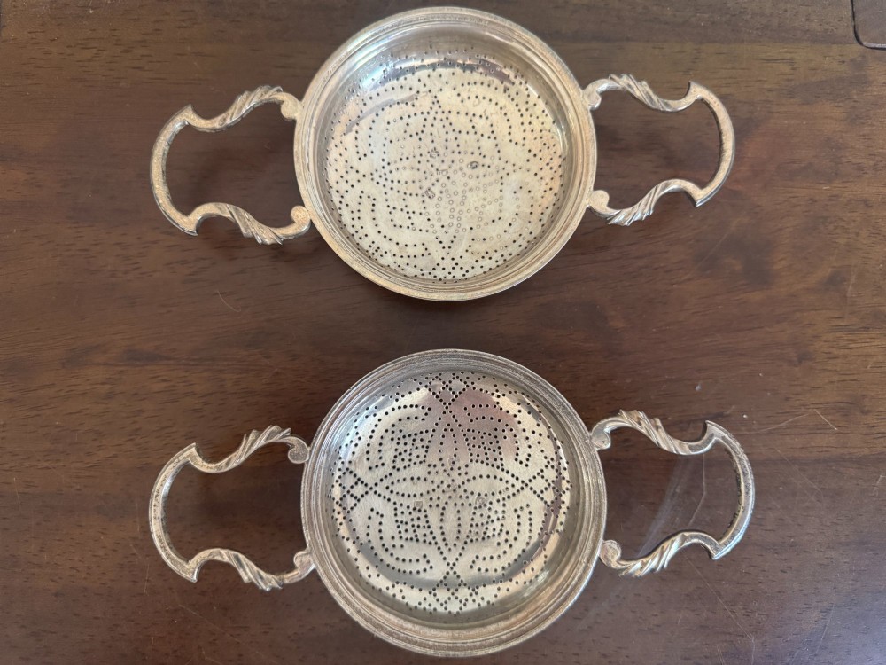 A matched pair of George III sterling silver lemon strainers featuring pierced geometric bowl patterns and dual side handles.