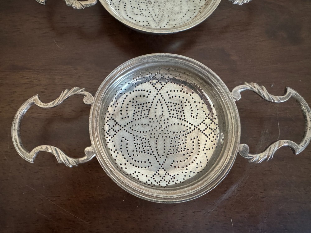 A matched pair of George III sterling silver lemon strainers featuring pierced geometric bowl patterns and dual side handles. View IV