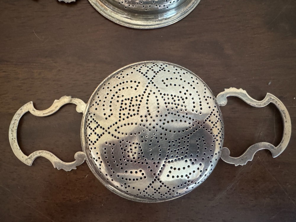 A matched pair of George III sterling silver lemon strainers featuring pierced geometric bowl patterns and dual side handles. View X