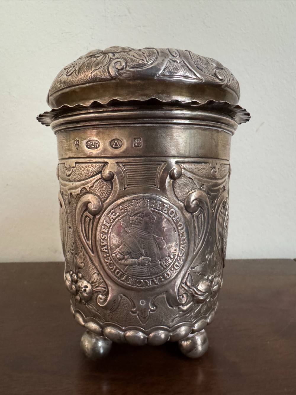 A sterling silver lidded beaker decorated with chased repousse scrollwork, embedded silver thaler coins, and standing on ball feet.