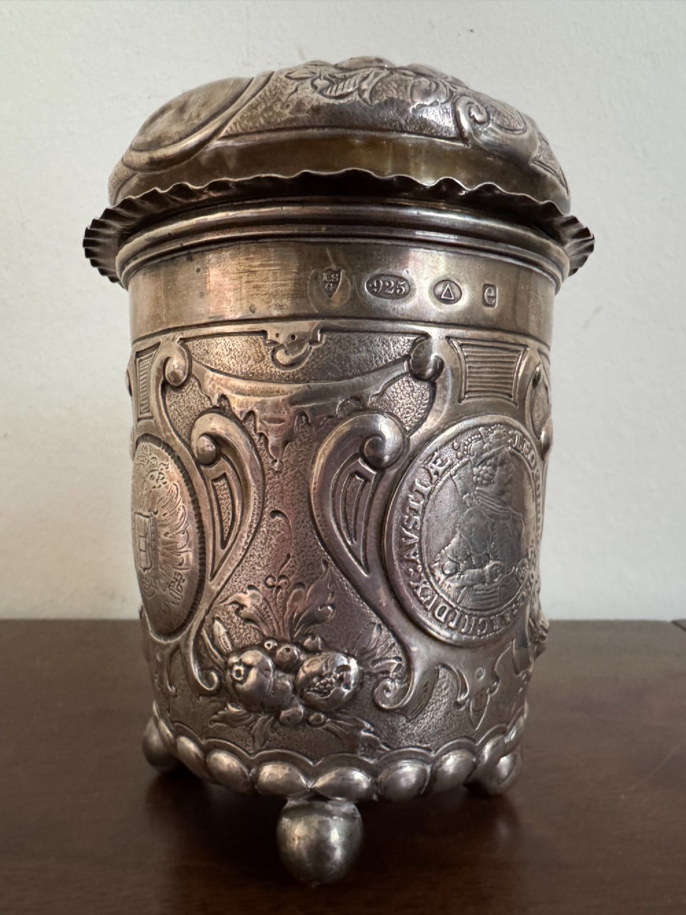 A sterling silver lidded beaker decorated with chased repousse scrollwork, embedded silver thaler coins, and standing on ball feet. View II