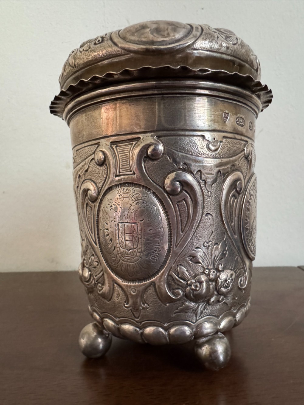A sterling silver lidded beaker decorated with chased repousse scrollwork, embedded silver thaler coins, and standing on ball feet. View III