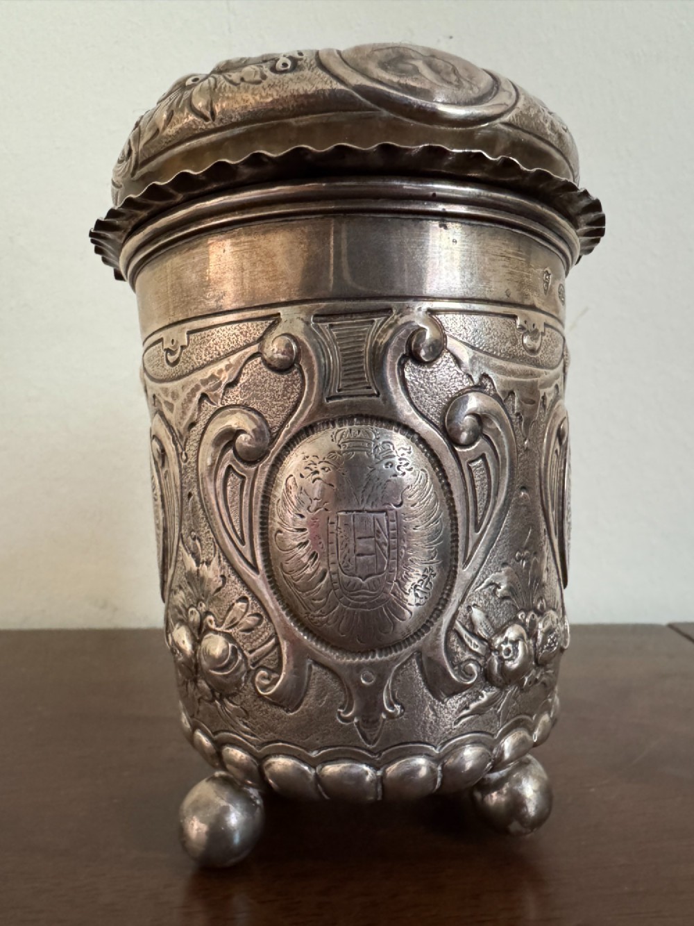 A sterling silver lidded beaker decorated with chased repousse scrollwork, embedded silver thaler coins, and standing on ball feet. View IV