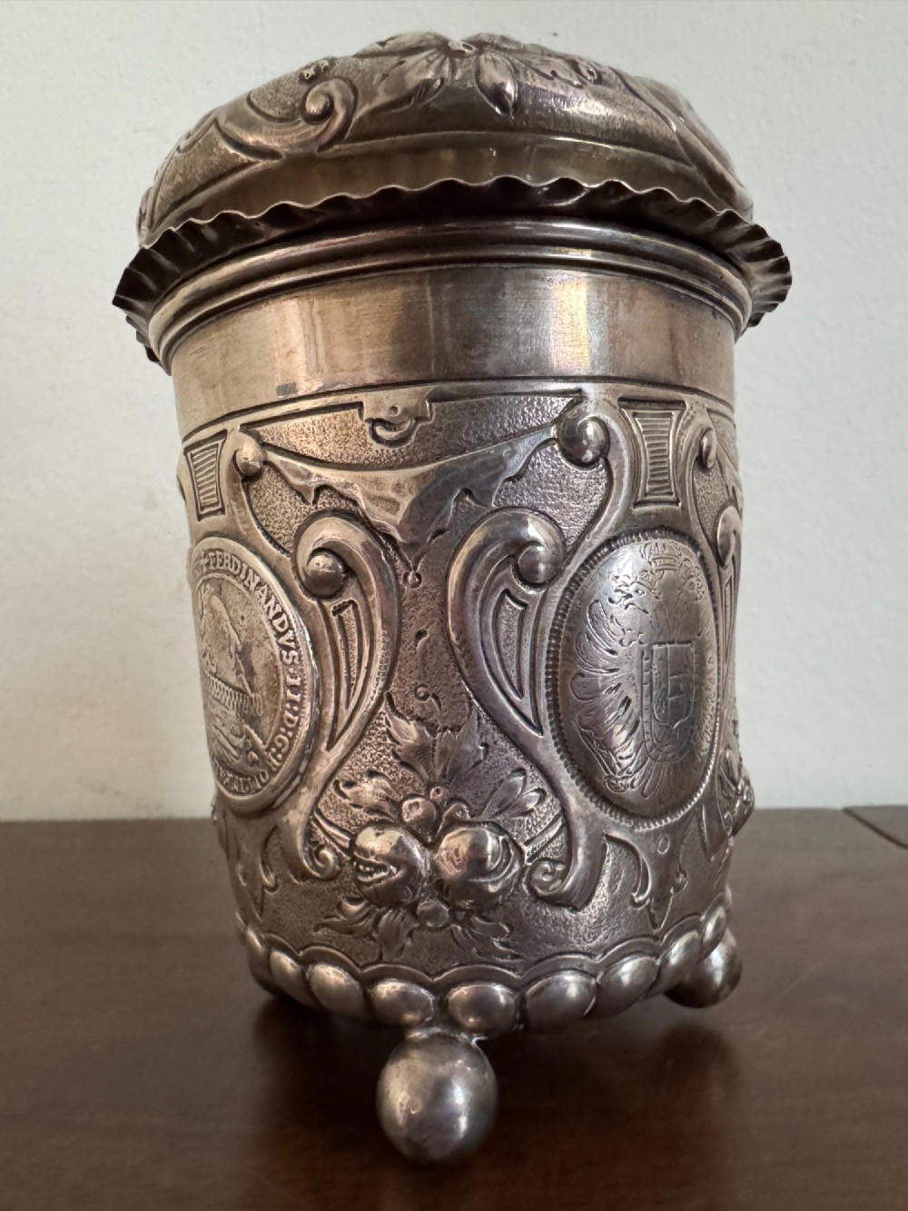A sterling silver lidded beaker decorated with chased repousse scrollwork, embedded silver thaler coins, and standing on ball feet. View V