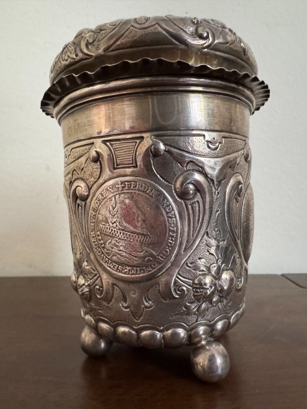 A sterling silver lidded beaker decorated with chased repousse scrollwork, embedded silver thaler coins, and standing on ball feet. View VI