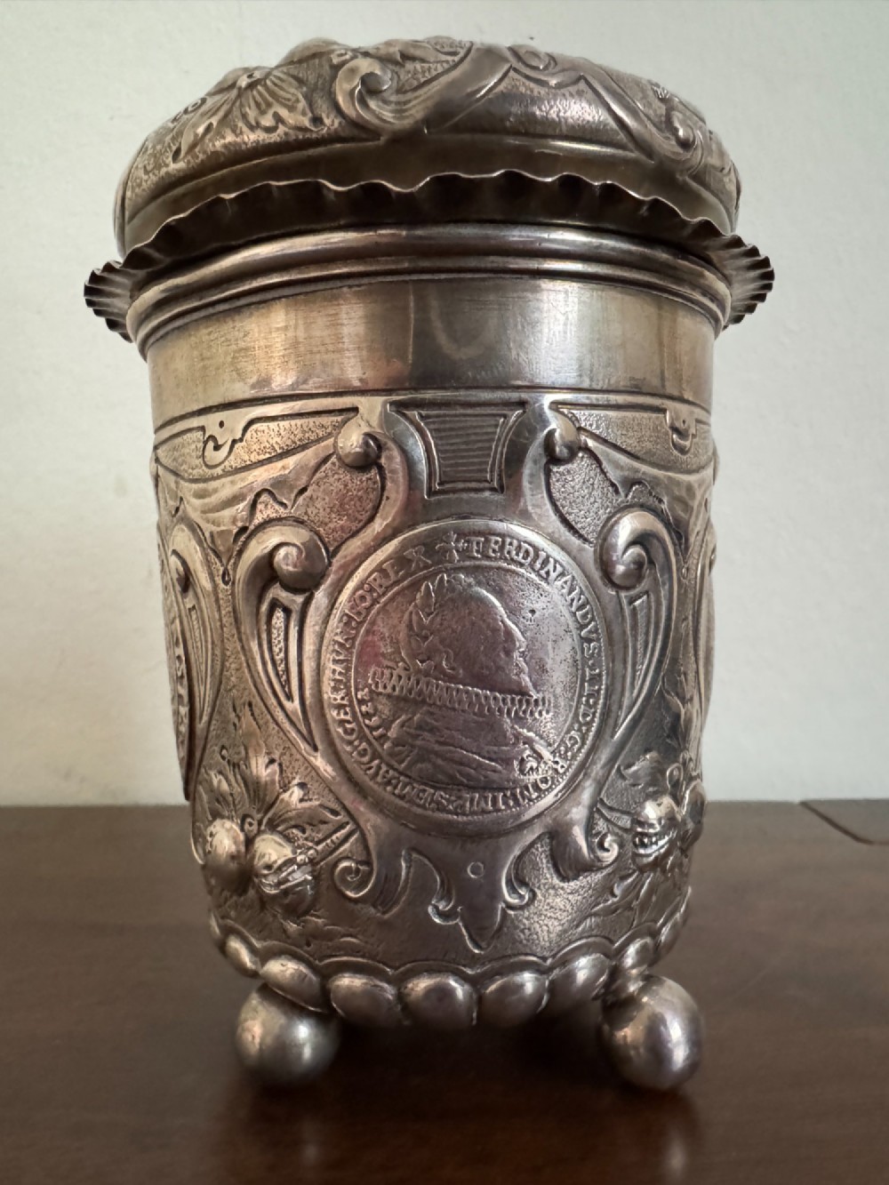 A sterling silver lidded beaker decorated with chased repousse scrollwork, embedded silver thaler coins, and standing on ball feet. View VII