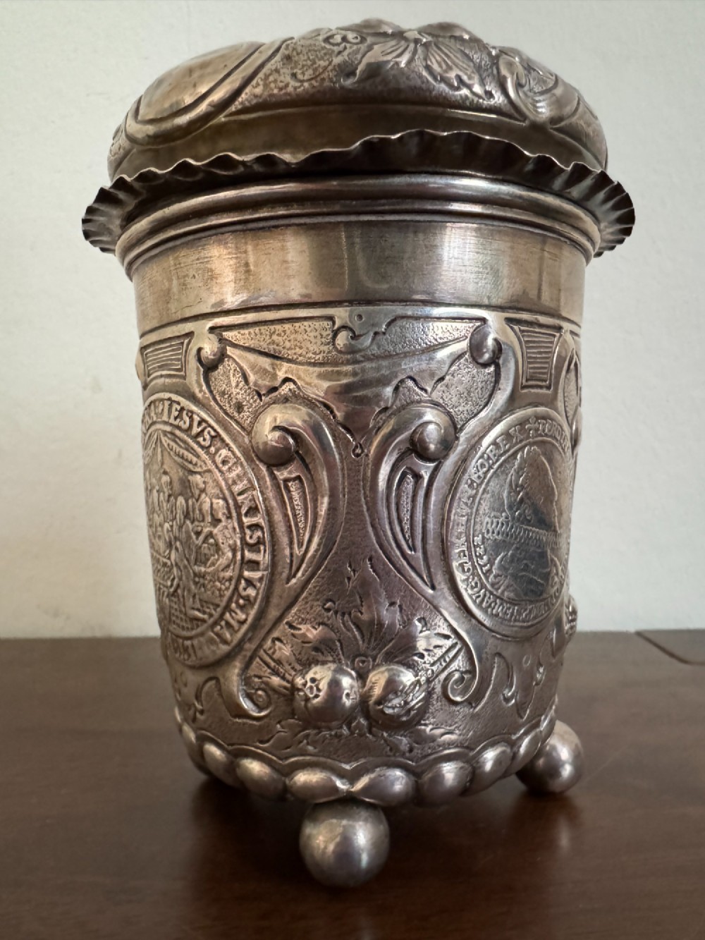 A sterling silver lidded beaker decorated with chased repousse scrollwork, embedded silver thaler coins, and standing on ball feet. View VIII