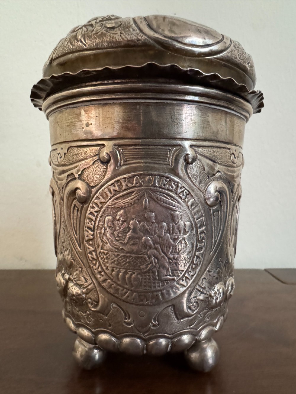 A sterling silver lidded beaker decorated with chased repousse scrollwork, embedded silver thaler coins, and standing on ball feet. View IX