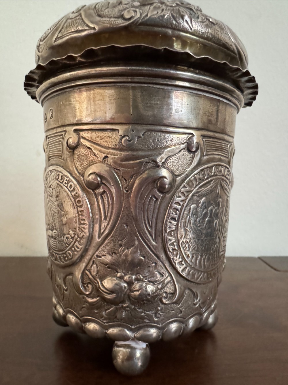 A sterling silver lidded beaker decorated with chased repousse scrollwork, embedded silver thaler coins, and standing on ball feet. View X