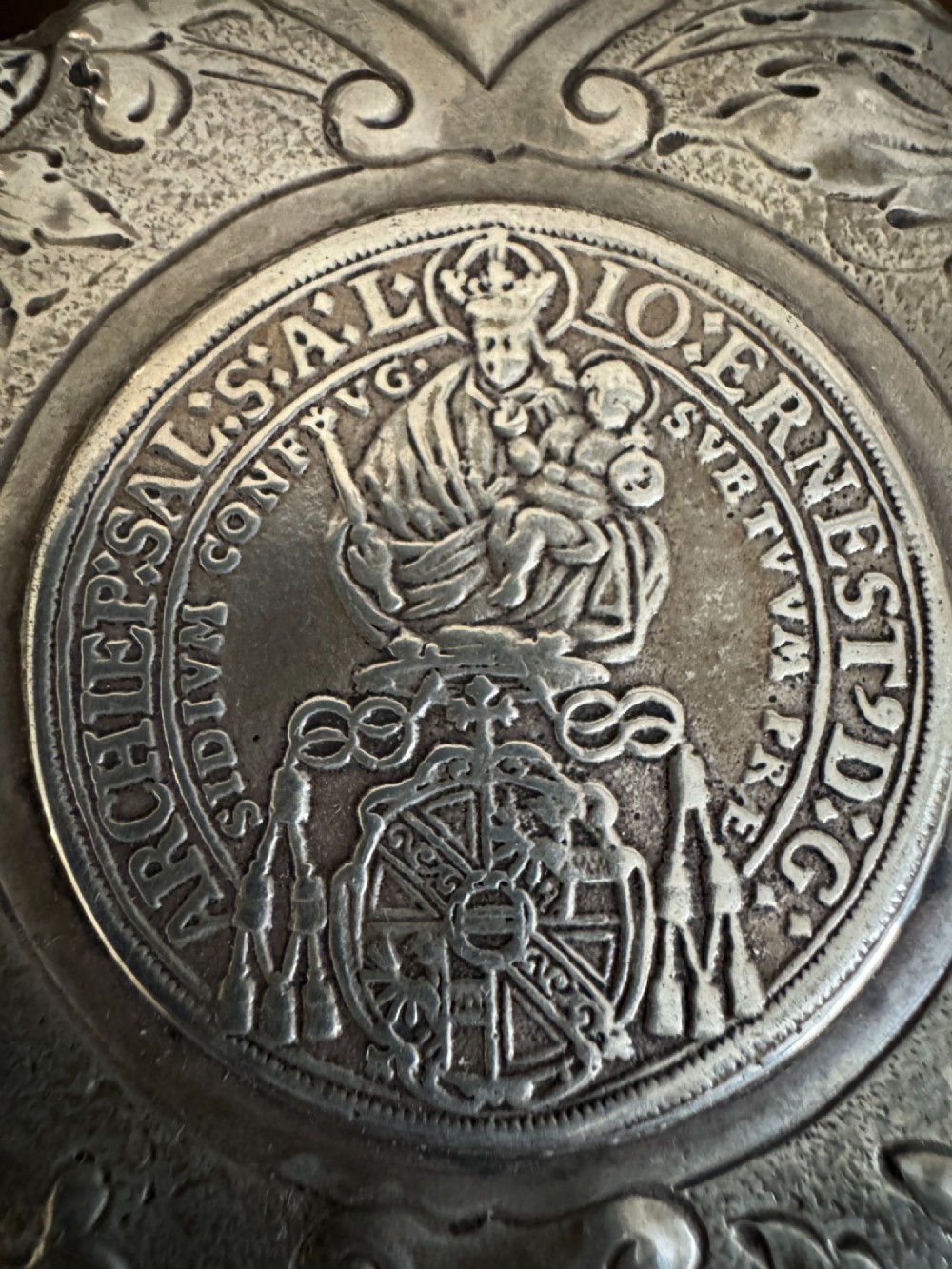 A sterling silver lidded beaker decorated with chased repousse scrollwork, embedded silver thaler coins, and standing on ball feet. View XV