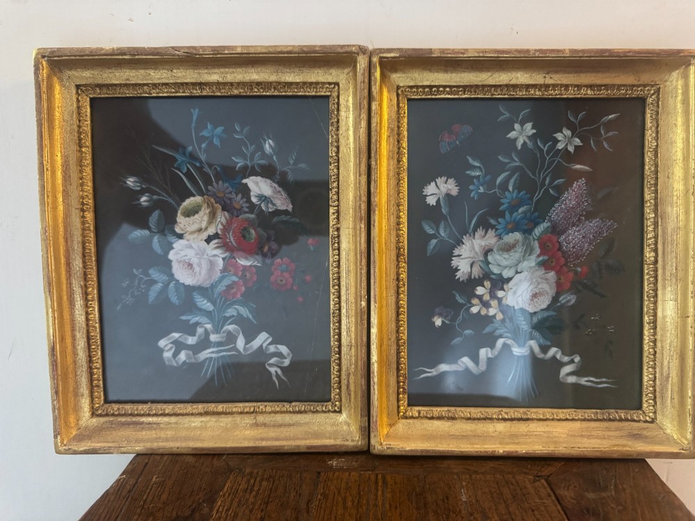 A pair of framed antique gouache paintings on dark backgrounds depicting floral bouquets tied with white ribbons, set in gilded frames.