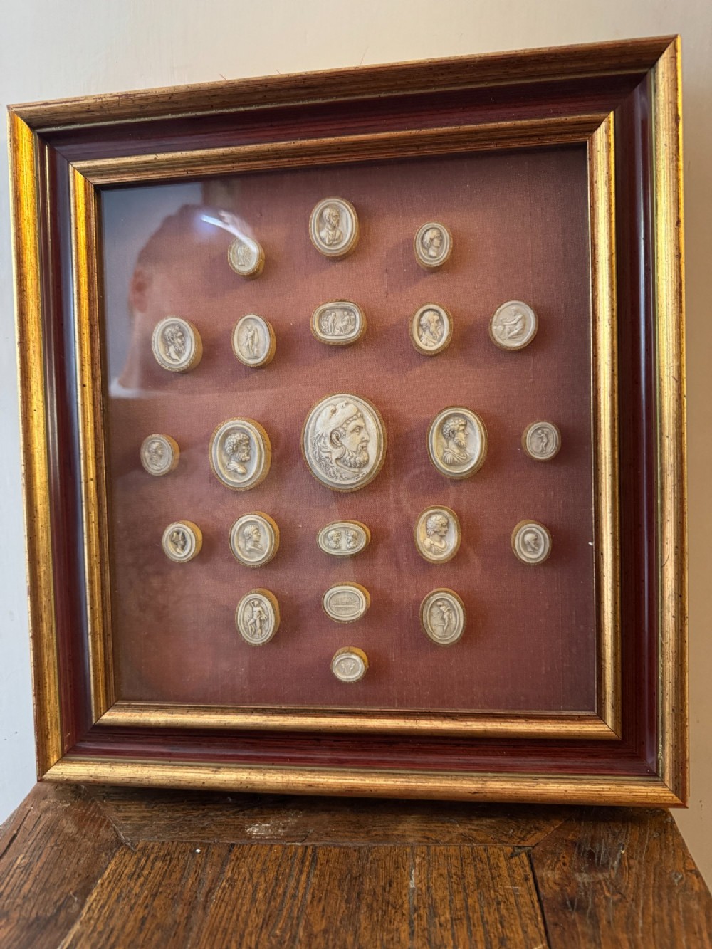 A square gilt and red frame containing nineteen oval plaster intaglio medallions symmetrically arranged on crimson velvet background.