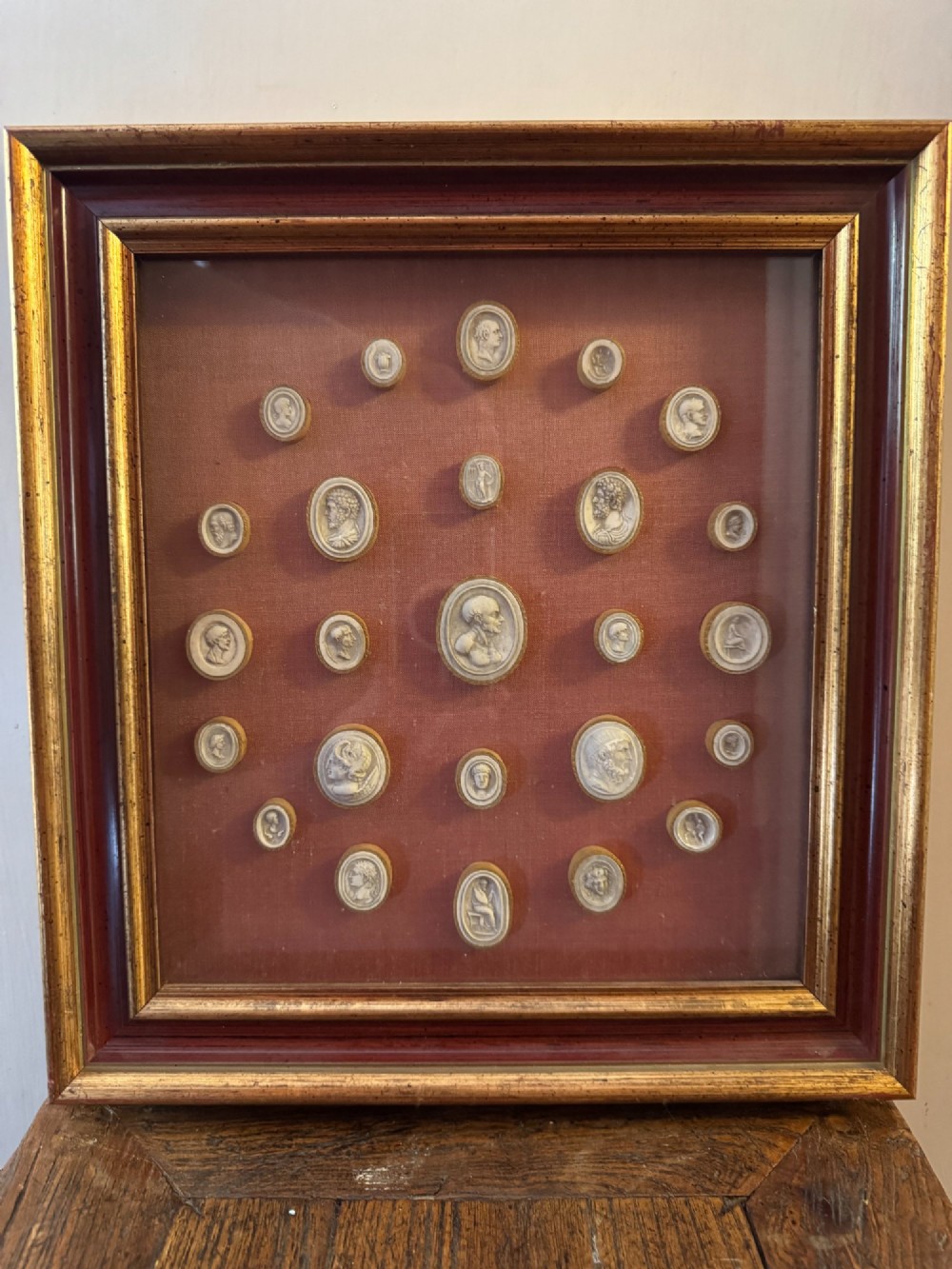 A square gilt and red frame containing nineteen oval plaster intaglio medallions symmetrically arranged on crimson velvet background. View IV