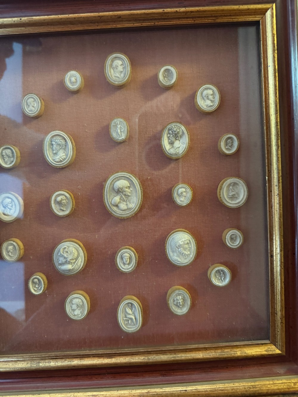 A square gilt and red frame containing nineteen oval plaster intaglio medallions symmetrically arranged on crimson velvet background. View VI