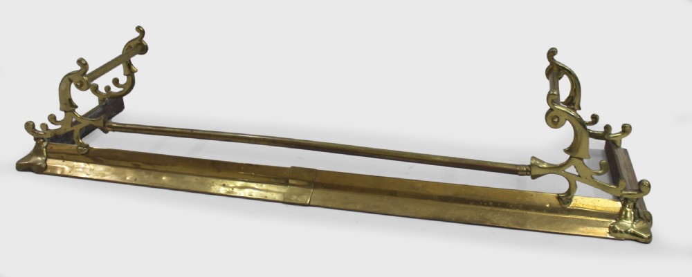 A Victorian brass fire fender with cast scroll end supports and a central horizontal rail resting on a low metal kerb.