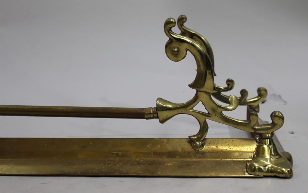 A Victorian brass fire fender with cast scroll end supports and a central horizontal rail resting on a low metal kerb. View III