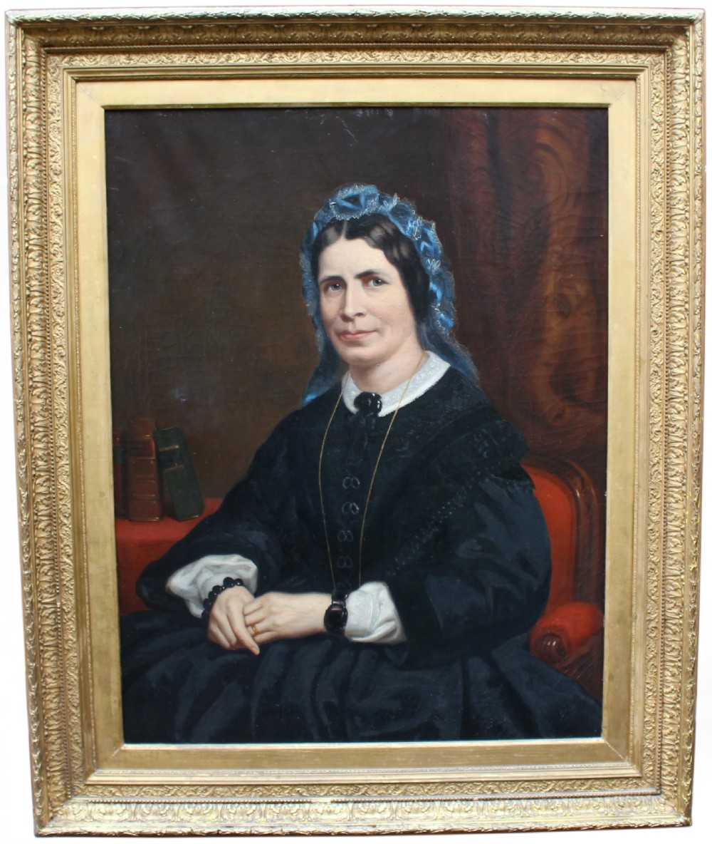 A mid-Victorian oil portrait of a seated lady wearing a black dress and blue lace bonnet inside a gilded wood frame.