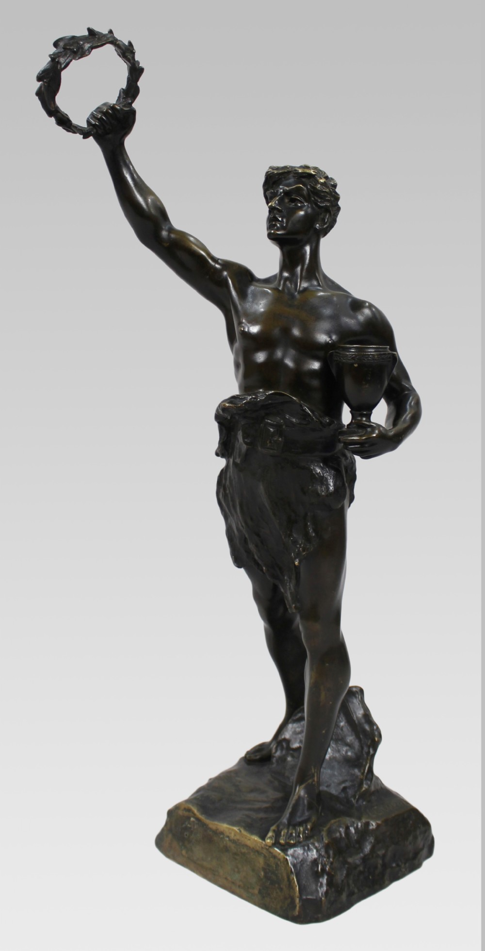 A dark patinated bronze sculpture depicting a standing classical male figure raising a laurel wreath in his right hand and holding a cup in his left hand.