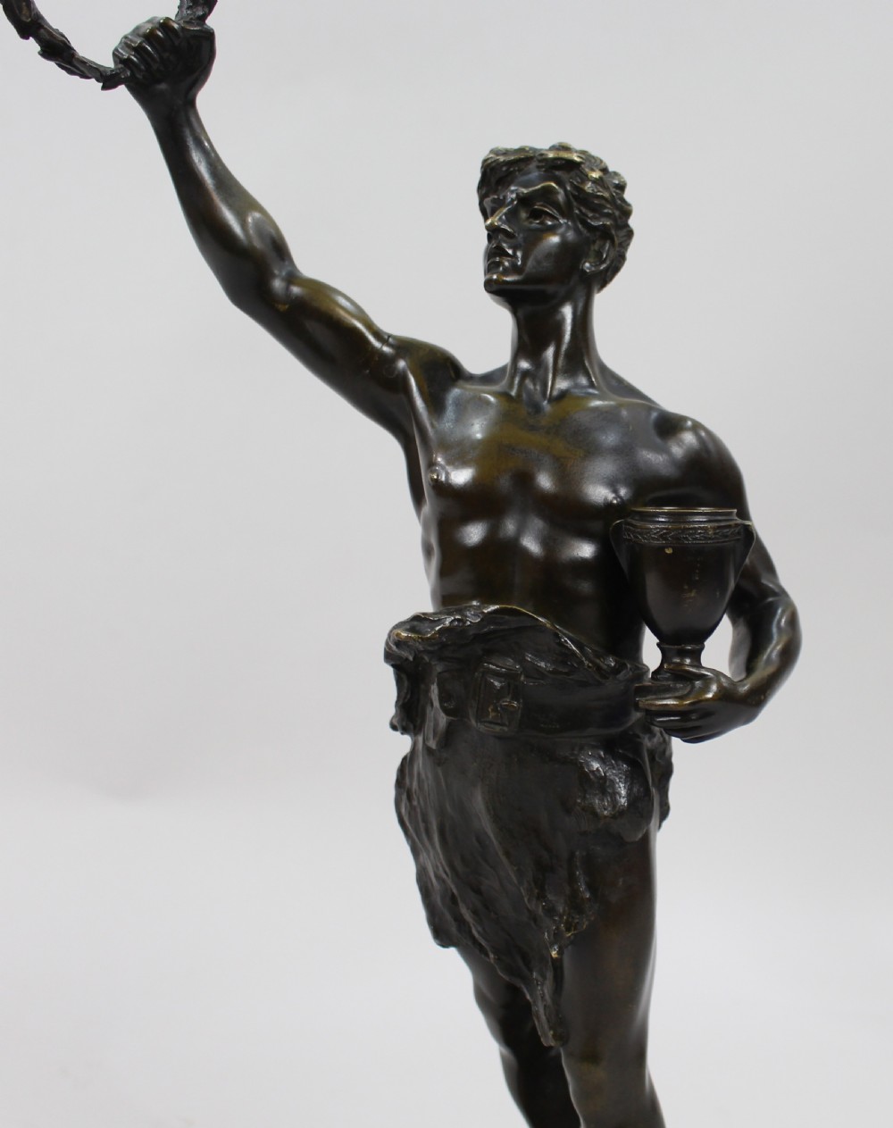 A dark patinated bronze sculpture depicting a standing classical male figure raising a laurel wreath in his right hand and holding a cup in his left hand. View XI