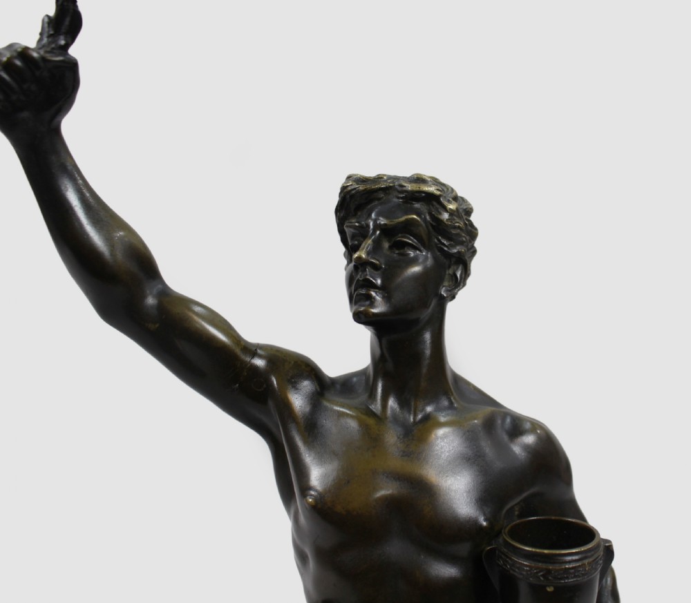 A dark patinated bronze sculpture depicting a standing classical male figure raising a laurel wreath in his right hand and holding a cup in his left hand. View XII