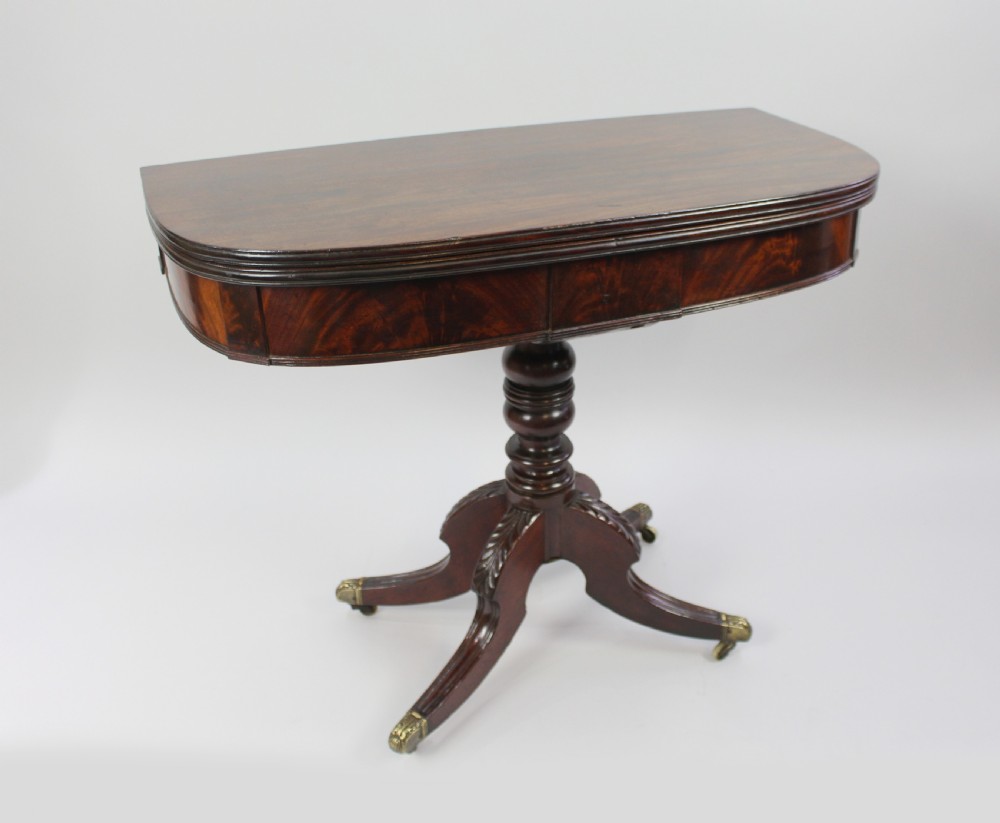 An early nineteenth-century Regency flame mahogany tea table featuring a fold-over top, turned central pedestal column, and four carved splayed legs with brass castors. View II