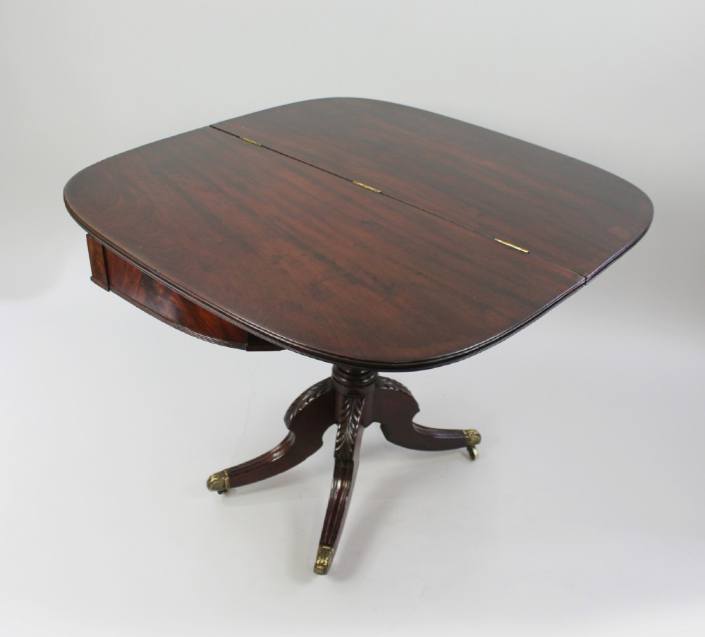 An early nineteenth-century Regency flame mahogany tea table featuring a fold-over top, turned central pedestal column, and four carved splayed legs with brass castors. View V