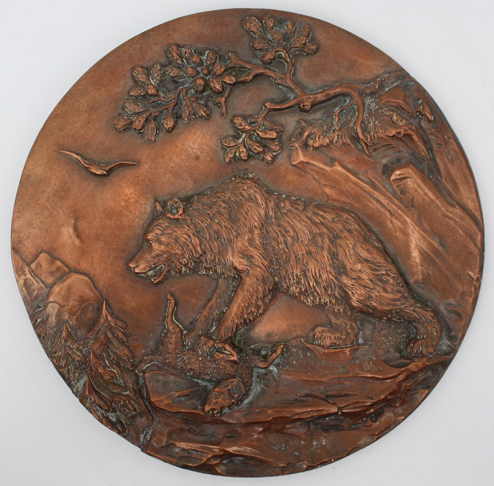 A circular antique copper relief plaque depicting a bear with prey in a rocky forest setting under an oak branch.