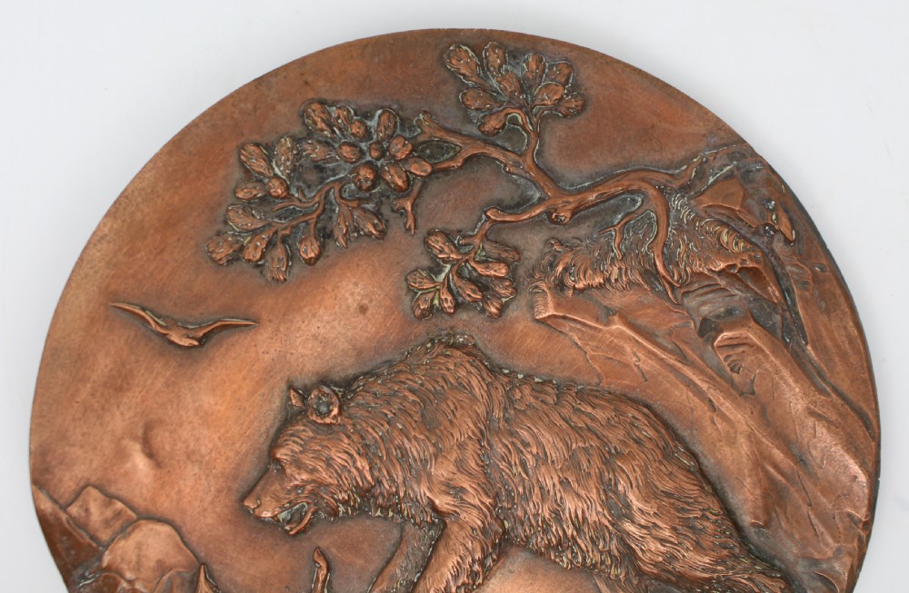 A circular antique copper relief plaque depicting a bear with prey in a rocky forest setting under an oak branch. View II