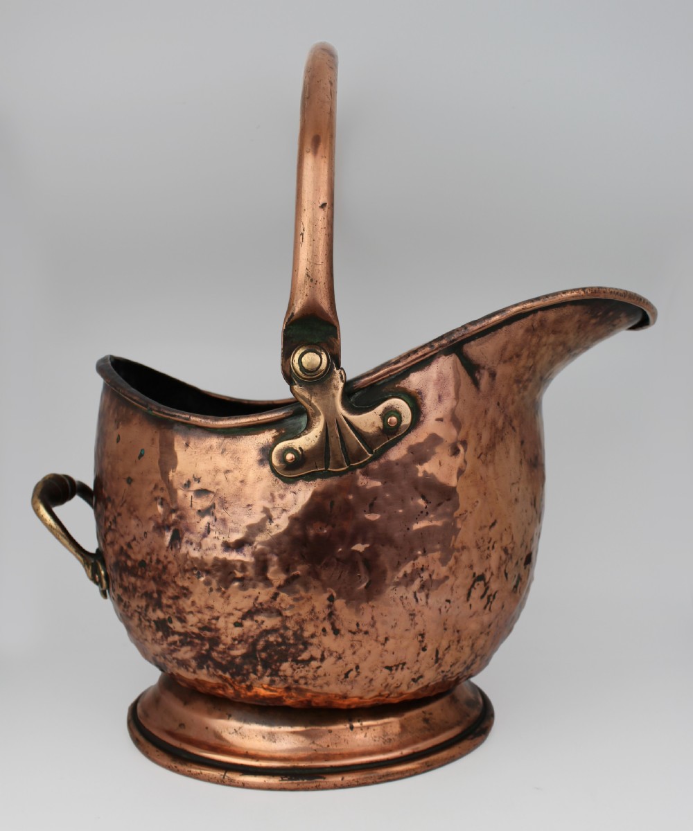 A Victorian helmet-shaped copper coal scuttle featuring an overhead swing handle, rear brass handle, and a round pedestal base.