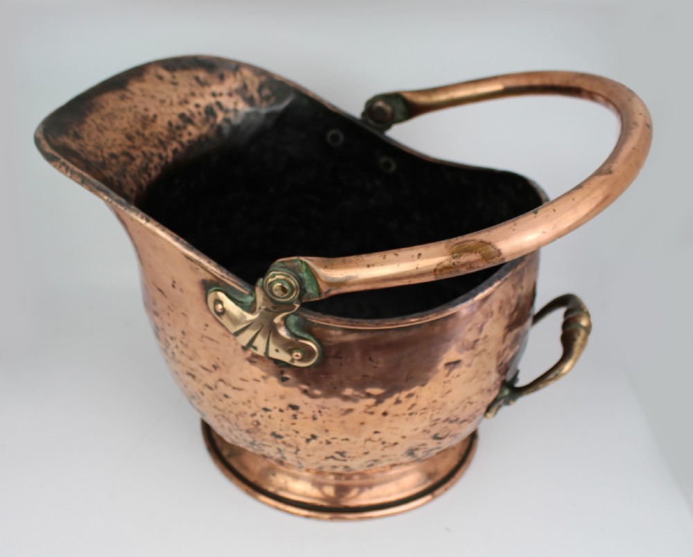 A Victorian helmet-shaped copper coal scuttle featuring an overhead swing handle, rear brass handle, and a round pedestal base. View V