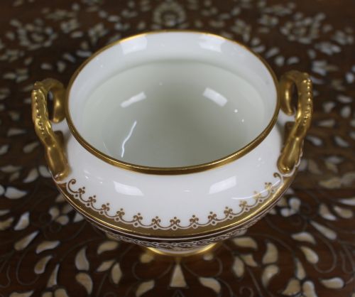 fig 1. A 19th-century white porcelain urn featuring gilded side handles, a classical scroll frieze, and a fluted pedestal foot.