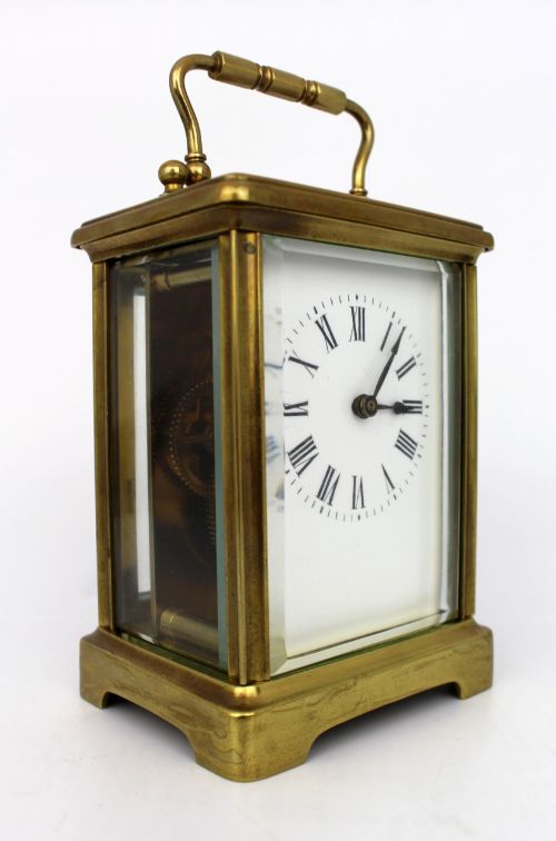 fig 1. An antique brass carriage clock featuring rectangular glass side panels, a white enamel dial with black Roman numerals, and a folding top handle.