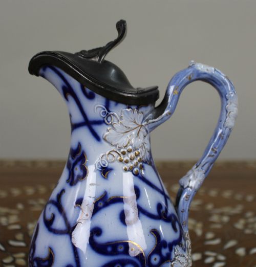 fig 1. A Victorian relief-moulded flow blue earthenware jug decorated with cobalt scrollwork, white floral details with gold accents, and a hinged pewter lid.
