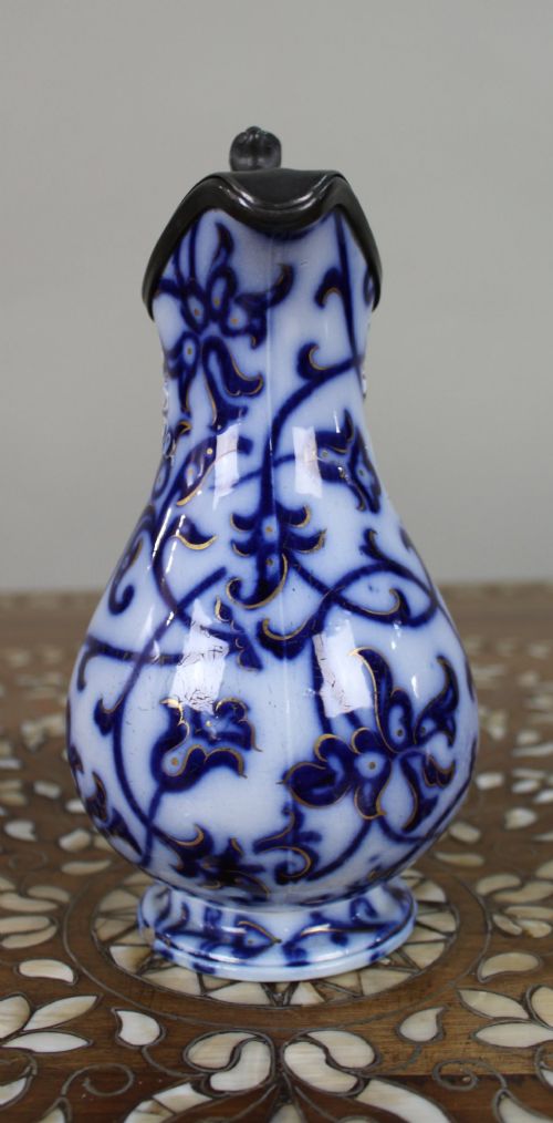 A Victorian relief-moulded flow blue earthenware jug decorated with cobalt scrollwork, white floral details with gold accents, and a hinged pewter lid. View IV