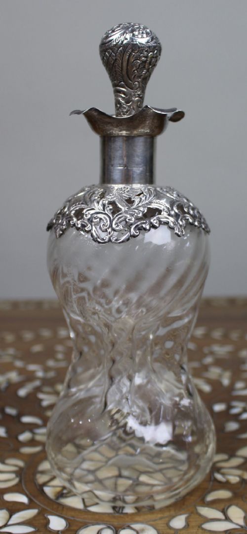 fig 1. An antique clear glass decanter featuring a pinched waist, swirled body, ornate pierced silver shoulder mount, silver collar, and a chased silver stopper.