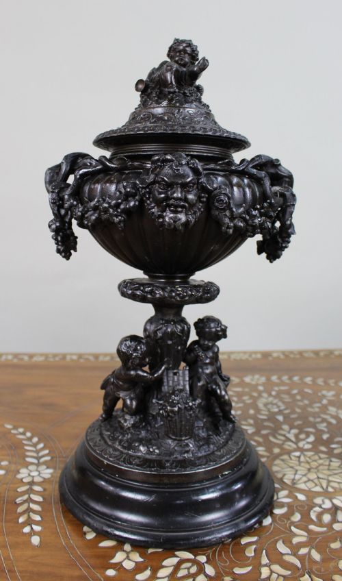 fig 1. A pair of dark bronzed spelter urns with lids topped by seated putti figures, featuring Bacchic masks, relief garlands, and bases supported by sculpted cherubs.