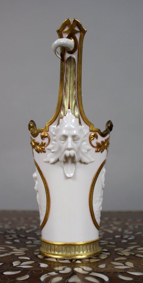 fig 1. A white porcelain vase featuring gilded scrollwork, dolphin handles, mascaron side mounts, and a central cameo medallion depicting a cherub.