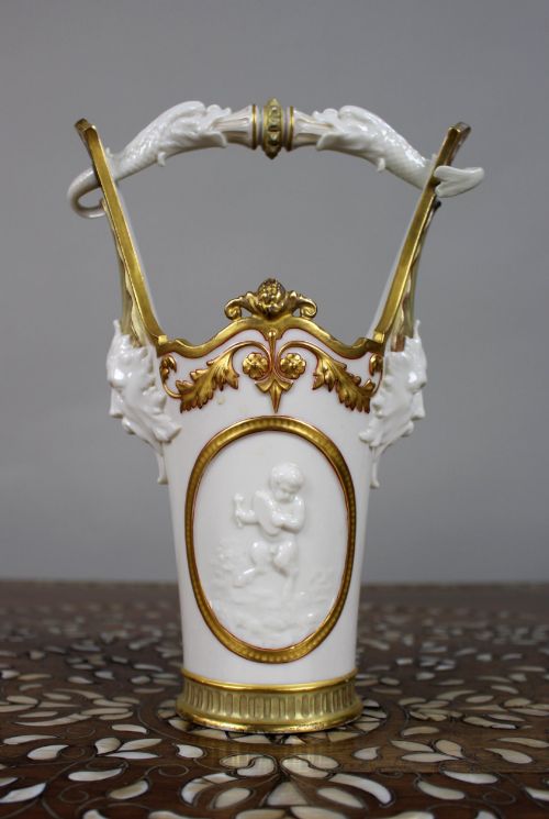 fig 2. A white porcelain vase featuring gilded scrollwork, dolphin handles, mascaron side mounts, and a central cameo medallion depicting a cherub.