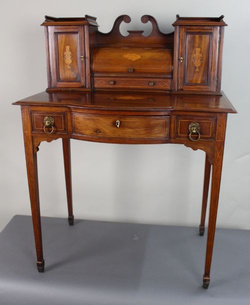 fig 1. An Edwardian inlaid mahogany writing desk featuring a superstructure with small side cabinets, shell motif marquetry, a bow-front central drawer, and square tapered legs.