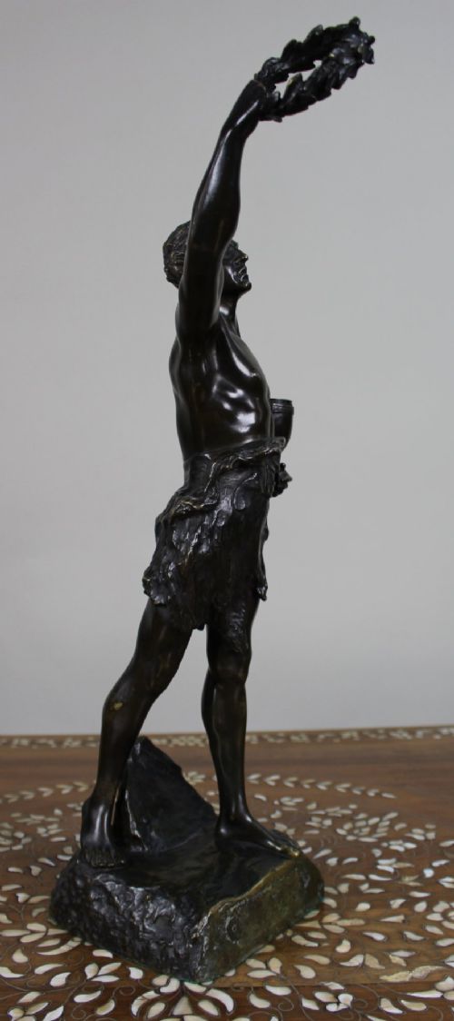 fig 1. A dark patinated bronze figure of a classical male holding a raised wreath in one hand and a vessel in the other.