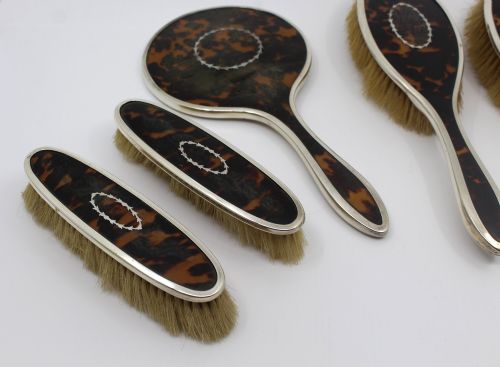 fig 1. A five-piece Art Deco sterling silver and faux tortoiseshell vanity set featuring a hand mirror, two handled hairbrushes, and two clothes brushes with silver leaf-wreath inlays.