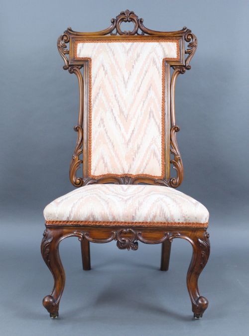 fig 1. A Victorian carved walnut occasional chair featuring a tall upholstered backrest with patterned beige fabric, scroll-carved cresting, cabriole legs, and ceramic castors.