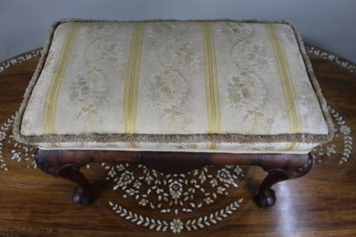 fig 1. A low walnut footstool featuring carved cabriole legs and a cream and yellow floral striped fabric upholstered seat with tassel trim.
