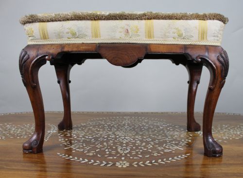fig 2. A low walnut footstool featuring carved cabriole legs and a cream and yellow floral striped fabric upholstered seat with tassel trim.