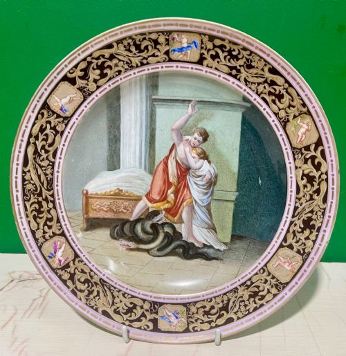 fig 2. A circular 19th-century Meissen porcelain cabinet plate featuring a central hand-painted classical scene framed by a dark burgundy rim with intricate gilded scrollwork and cherub cartouches.