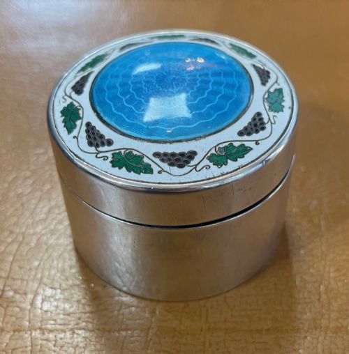 fig 1. A circular Edwardian silver box lid featuring a blue engine-turned guilloche enamel centre surrounded by a white enamel border with green foliage and dark grape motifs.
