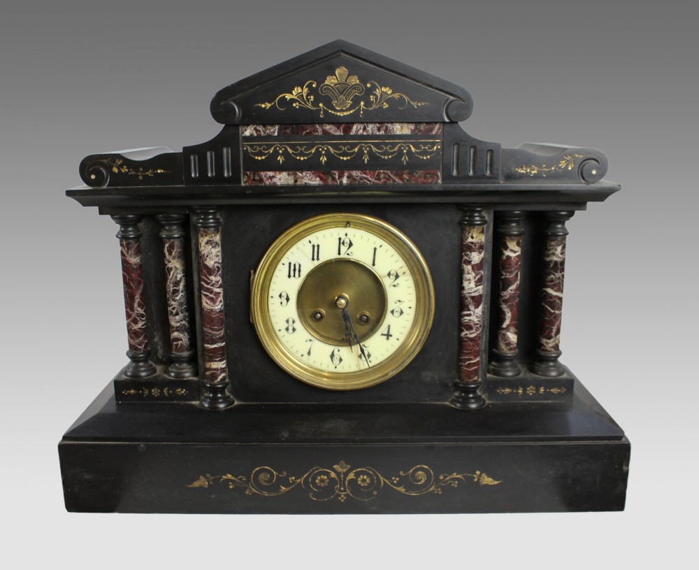 A High Victorian architectural mantel clock crafted from black slate with red rouge marble columns, a brass dial, and gilt floral engravings.
