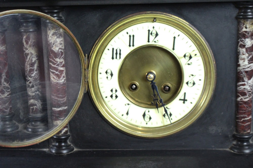 A High Victorian architectural mantel clock crafted from black slate with red rouge marble columns, a brass dial, and gilt floral engravings. View V