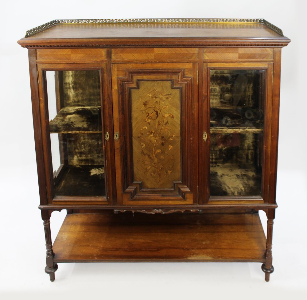 Late Victorian rosewood display cabinet featuring a central inlaid floral door, two glass side doors, a brass gallery rail top, and a lower tier shelf on turned legs.
