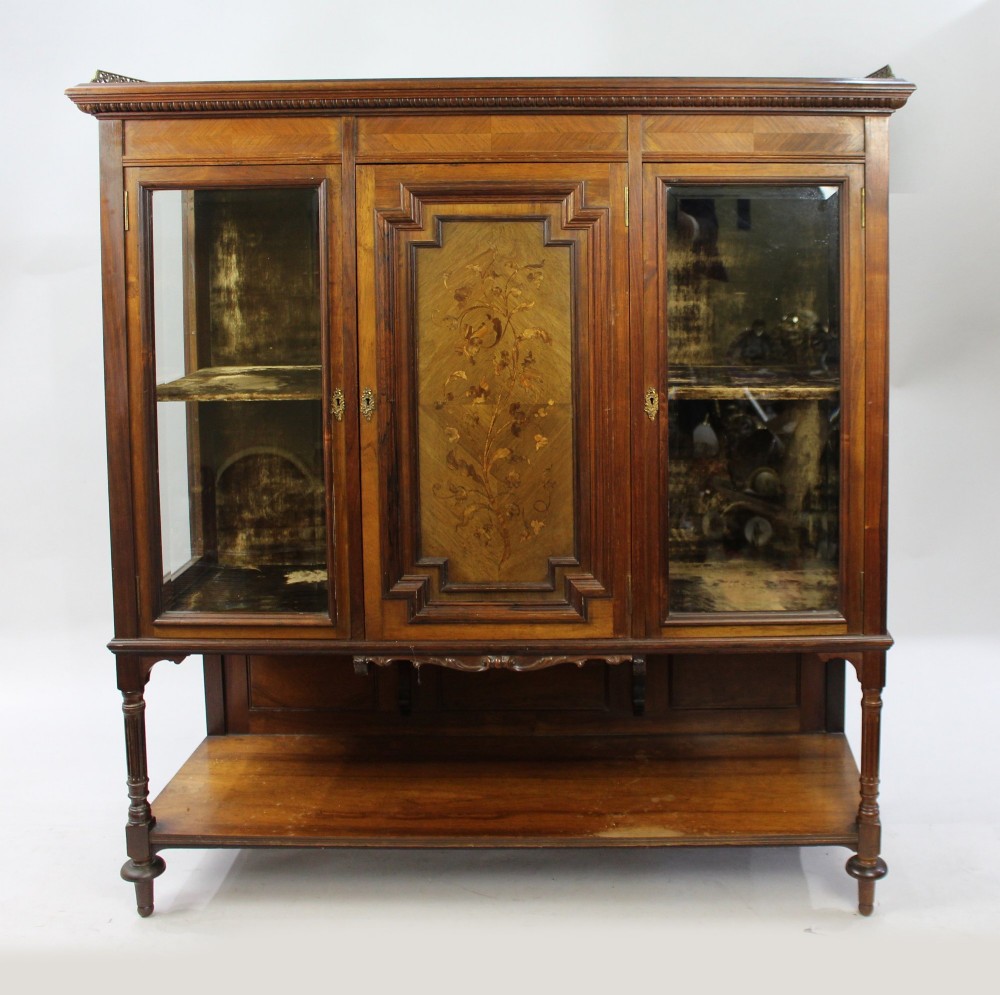 Late Victorian rosewood display cabinet featuring a central inlaid floral door, two glass side doors, a brass gallery rail top, and a lower tier shelf on turned legs. View II